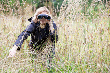 Naklejka na ściany i meble Blond hair European woman spying with binoculars, move to the side high dry grass, black jacket