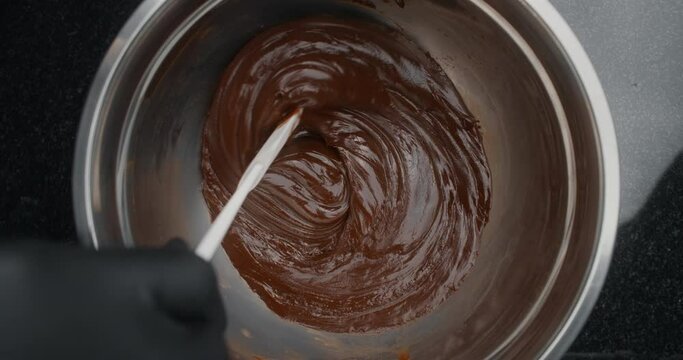 Tabletop shot: chef slowly stirs melted chocolate in metal bowl over low heat in slow motion, raw materials for making sweets and chocolate, cooking the sweet desserts, making chocolate candies, 4k
