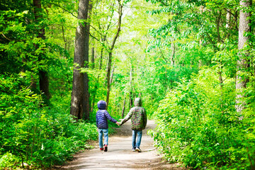 Two children in jackets are walking around city park or forest on a safe distance in a sunny day holding hands