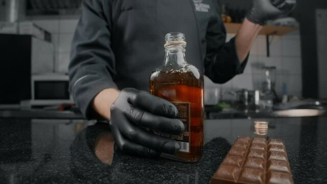 Zooming shot: chef in black apron and gloves opens the bottle of cognac and bung flies away in slow motion, opening the bottle of alcohol drink, splashes of whiskey, tabletop food video, Full HD 240