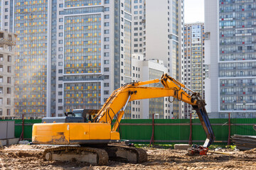 Obraz premium Heavy excavator in a working high frequency hydraulic vibratory mounted pile driver on road against the background of residential multi-storey buildings.