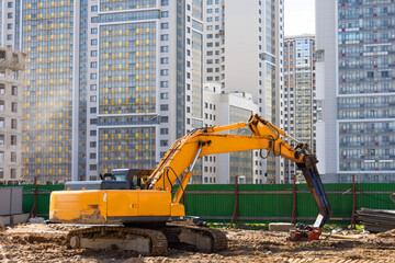 Heavy excavator in a working high frequency hydraulic vibratory mounted pile driver on road against the background of residential multi-storey buildings.