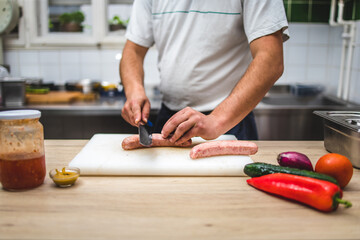 Male chef cutting raw meat sausage on the board with vegetables on the side.