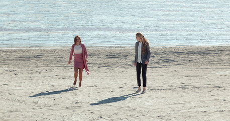 two women in office clothes run along the sand of the sea shore.
