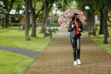 A masked girl is walking along the street. A girl in a protective mask walks in the park with an umbrella in the rain. Coronavirus infection COVID-19