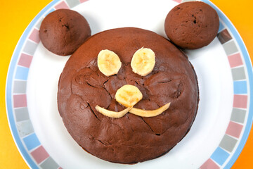 Homemade chocolate cake with eyes, nose and mouth made from banana. Smiling face of brown bear. Chocolate cupcakes on plate in shape of bear head