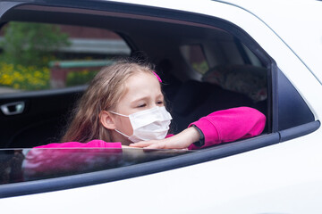 a caucasian little  girl in a medical mask looks out the open window of a white car. She put her head in her hands. Hygiene mask protection coronavirus or covid-19. Allergy to plants. Air pollution.