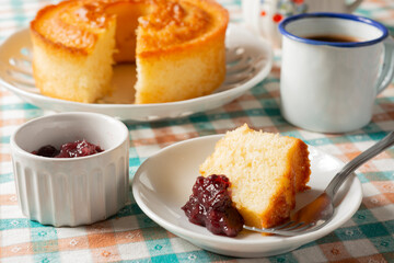 Cake, sliced cake, berry jam and a cup of coffee over plaid textile. Home breakfast scene.