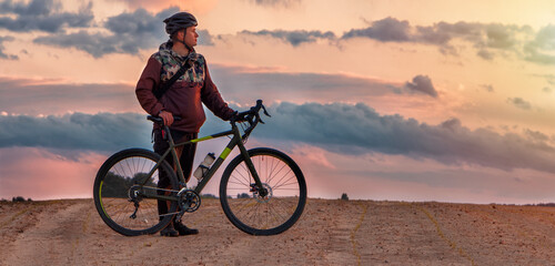 Guy in a helmet stands with a gravel bike in a sand field against a cloudy sky at sunset.