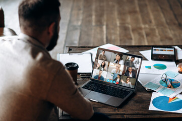 App for communication with employees. Online video conference. Young bearded man communicates via video call with colleagues, he sits at the creative modern office space - obrazy, fototapety, plakaty