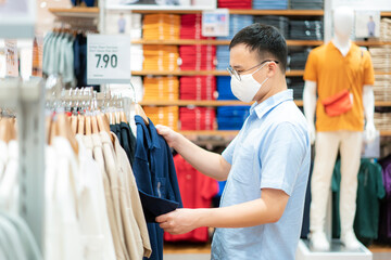 Asian man wearing mask over her face while choosing shirt at shopping mall with shopping bag for healthcare and prevention from coronavirus, Covid19 influenza in crowded place..
