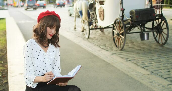 Portrait of beautiful charming paris girl in red beret drawing with pencil in notebook outdoor. Joyful pretty Caucasian woman resting while sitting in city and drawing in planner. Leisure concept