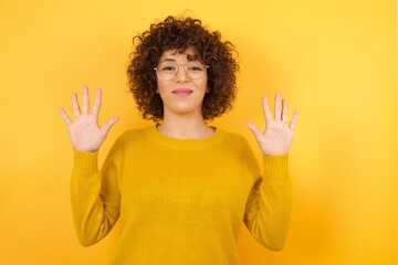 Young beautiful with curly hair wearing yellow sweater standing over isolated yellow background showing and pointing up with fingers number ten while smiling confident and happy.