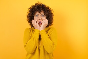 Photo of fearful Arab woman keeps hands near mouth, feels frightened and scared,  has a phobia, wearing yellow sweater, has Afro hairstyle. Shock and frighted concept.