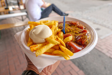 Close up view, Hand hold Currywurst, grilled pork sausage on top with curry powder served with curry ketchup sauce, Mayonnaise and Fried potatoes on plastic white plate and background of street. 