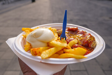 Close up view, Hand hold Currywurst, grilled pork sausage on top with curry powder served with curry ketchup sauce, Mayonnaise and Fried potatoes on plastic white plate and background of street. 