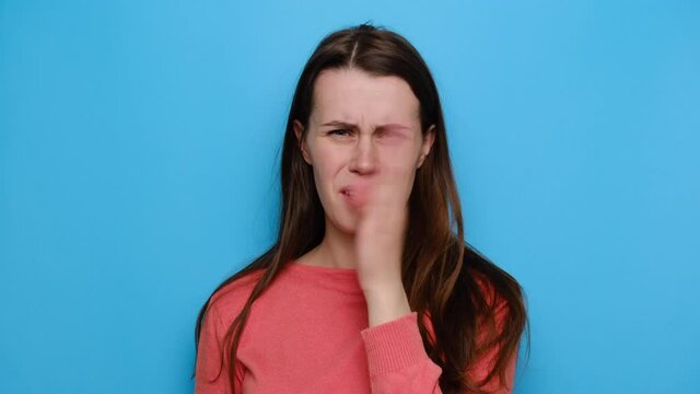 Close up portrait of annoyed young brunette woman checking her breath, smells something awful, pinches nose, dressed in sweater, isolated over blue studio background. Bad odour concept.