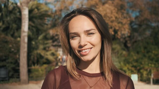 Tracking shot of beautiful romantic girl with beautiful cute smile charmingly looking in camera in city park. Attractive young woman with blue eyes