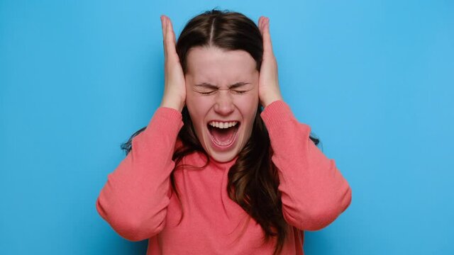 Portrait of sad disappointed young woman grabbing her head and screaming in confusion or fear, wears red sweater, isolated over blue studio background. Negative human emotions and feelings concept