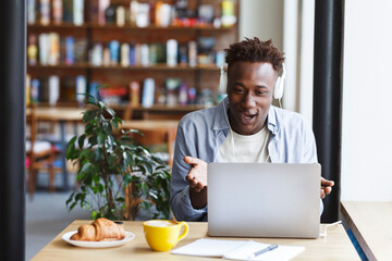 Excited African American guy with headset talking to his friends on video chat at coffee shop, blank space