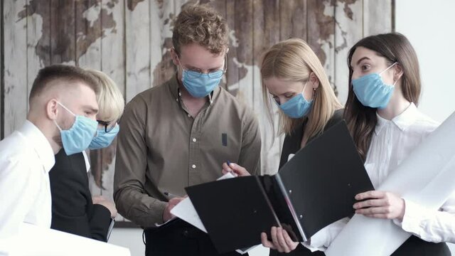 A Group Of Office Workers Are Standing Together In Masks. The Business Team Is Working On A Project In Masks As It Is COVID-19 Period. They Are Having Folders With Documents To Check.