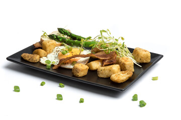 a fried egg sunny side up on a black plate with potatoes bacon asparagus and bread isolated on white
