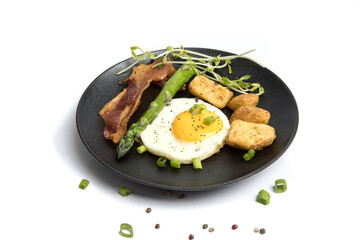 a fried egg sunny side up on a black plate with potatoes bacon asparagus and bread isolated on white
