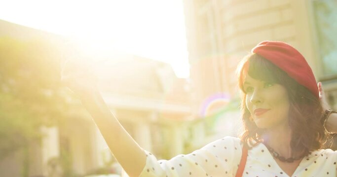 Close up portrait of pretty Caucasian young woman making selfie photo on mobile phone outdoor. Side view of beautiful girl taking pictures on smartphone in sunny light while standing on street.