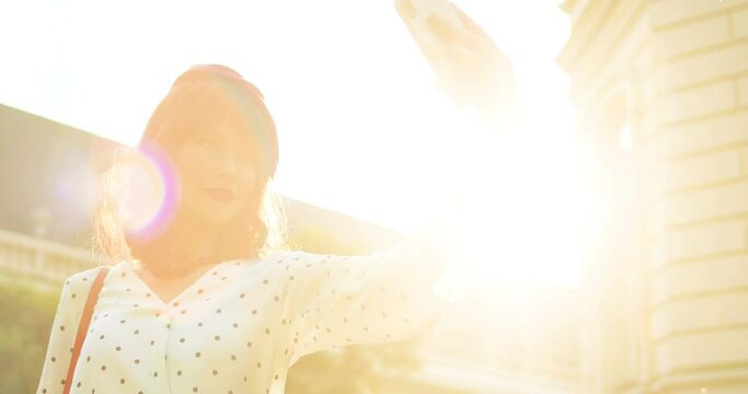 Portrait of pretty Caucasian young woman making selfie photo on mobile phone outdoor. Beautiful girl taking pictures on smartphone in sunny light while standing on street. Silhouette in sunshine