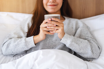A beautiful asian woman drinking hot coffee while lying on a white cozy bed at home in the morning