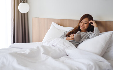 Obraz na płótnie Canvas A beautiful asian woman drinking hot coffee while lying on a white cozy bed at home in the morning