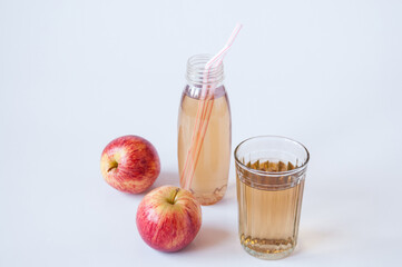 A bottle with straws and a glass of Apple juice and two red apples on a white background