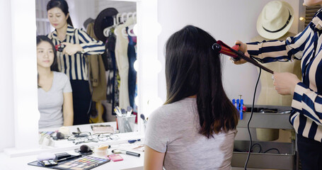 skilled young asian chinese stylist wrap curling hair in dressing room. girl actress getting ready before performance on backstage doing hairstyle. woman entertainer sit looking in mirror reflection