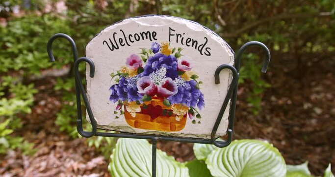 Beautiful, Welcome Friends slate sign Springtime floral garden in metal wire stand, start static then slow slide left against soft focus fluttering green hosta leaves in background