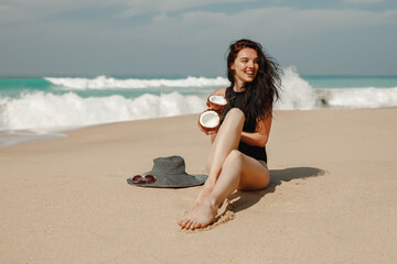 Young woman in  black swimsuit with coconut cocktail sit on the beach, ocean on background. Holding in hands two part of coconut and smiling. Summer vacation by the sea