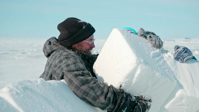 group of tourists builds igloo with large snow blocks and cut with metal saw on endless white frozen sea on sunny day