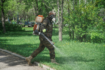 A worker in a dark protective suit conducts sanitary and chemical treatment in a garden or park. Spraying repellent or antiviral composition among green grass and trees