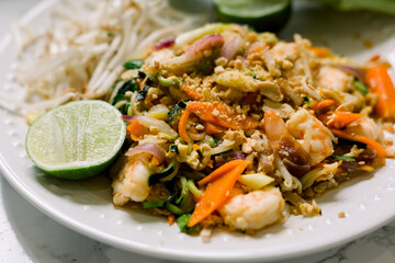 A close-up of shrimp pad thai cooked with carrots, zucchini noodles, bean sprouts, lime wedges, and crushed peanuts. 