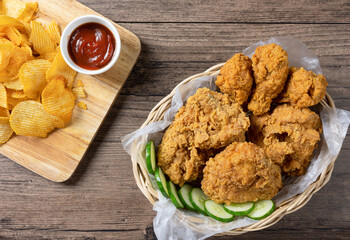 Crispy fried chicken in wicker basket serve with cucumber and potato chips on Wood cutting board serve with ketchup on wooden background. top view. Flat lay.