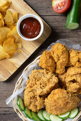 Crispy fried chicken in wicker basket serve with cucumber and potato chips on Wood cutting board serve with ketchup on wooden background. top view. Flat lay.