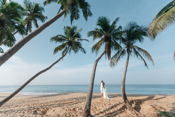 Happy newlyweds stand holding hands on the background of the blue sea and coconut palm trees Wedding walk on a sand beach