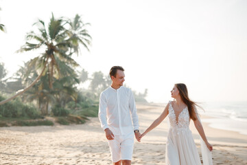 The bride and groom caucasian couple walk together walk along the beach sand shore. Coconut palm trees on background and ocean seascape. Honeymoon in asia