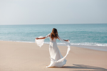 Fototapeta na wymiar Bride wearing white wedding dress spins, dancing ], plays wedding dress on white sand beach by the sea. Honeymoon near ocean