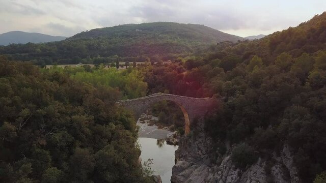 Aerial drone shot flying forward approaching a romanic bridge crossing a river surrounded by trees and vegetation. Sunset. Sadernes, Spain.