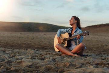 Young caucasian woman in white shirt and jean jacket playing guitar on the beach at the sun set.