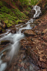 Obraz premium Majestic waterfall amid the dense lush green, with flowing stream and boulders in the foreground. Blurred effect on water. Carpathian, Ukraine