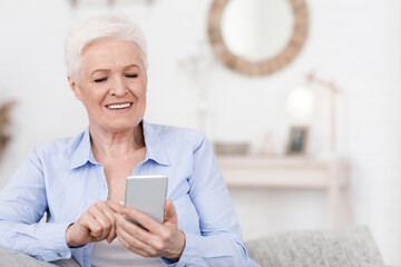 Smiling elderly woman holding cellphone, reading news while resting at home