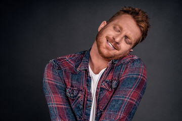 Silly and funny caucasian redhead guy with bristle, tilting head and leaning on shoulder with lovely and cute expression, smiling with closed eyes being tender and girly over gray background