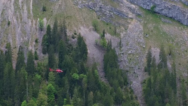 Red helicopter flying in german alps from left to right. Transport helicopter to restock remote lodged with new supplies. Mountain covered in forest in background