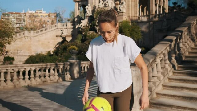 Tracking in shot of beautiful sporty girl doing kick ups with football and confidently looking in camera during training outdoor. Gorgeous girl doing football tricks